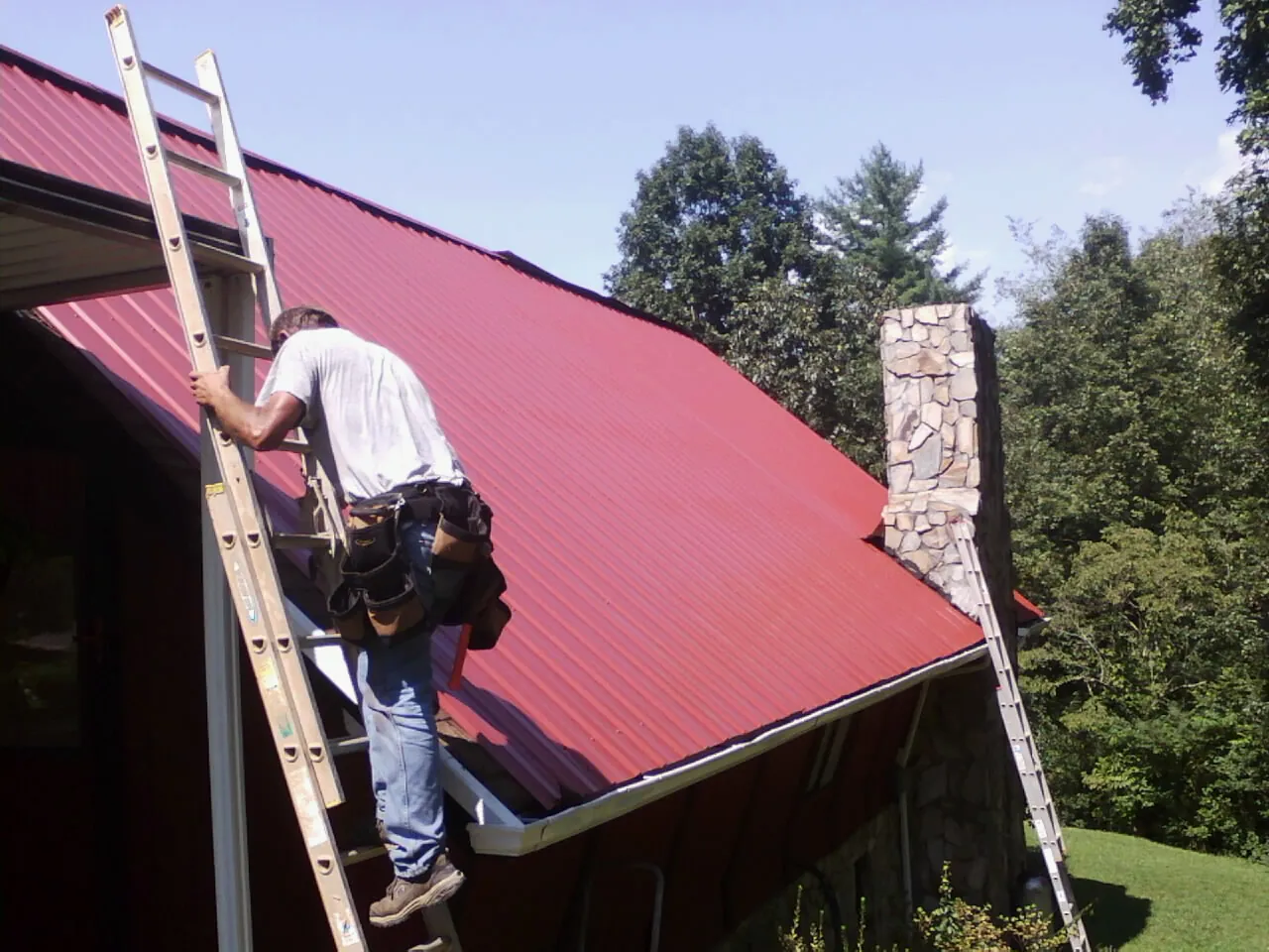 Roofer on a ladder beside a red metal roof and stone chimney — Soffit & Fascia Repair by Roofing Brothers in Larchmont